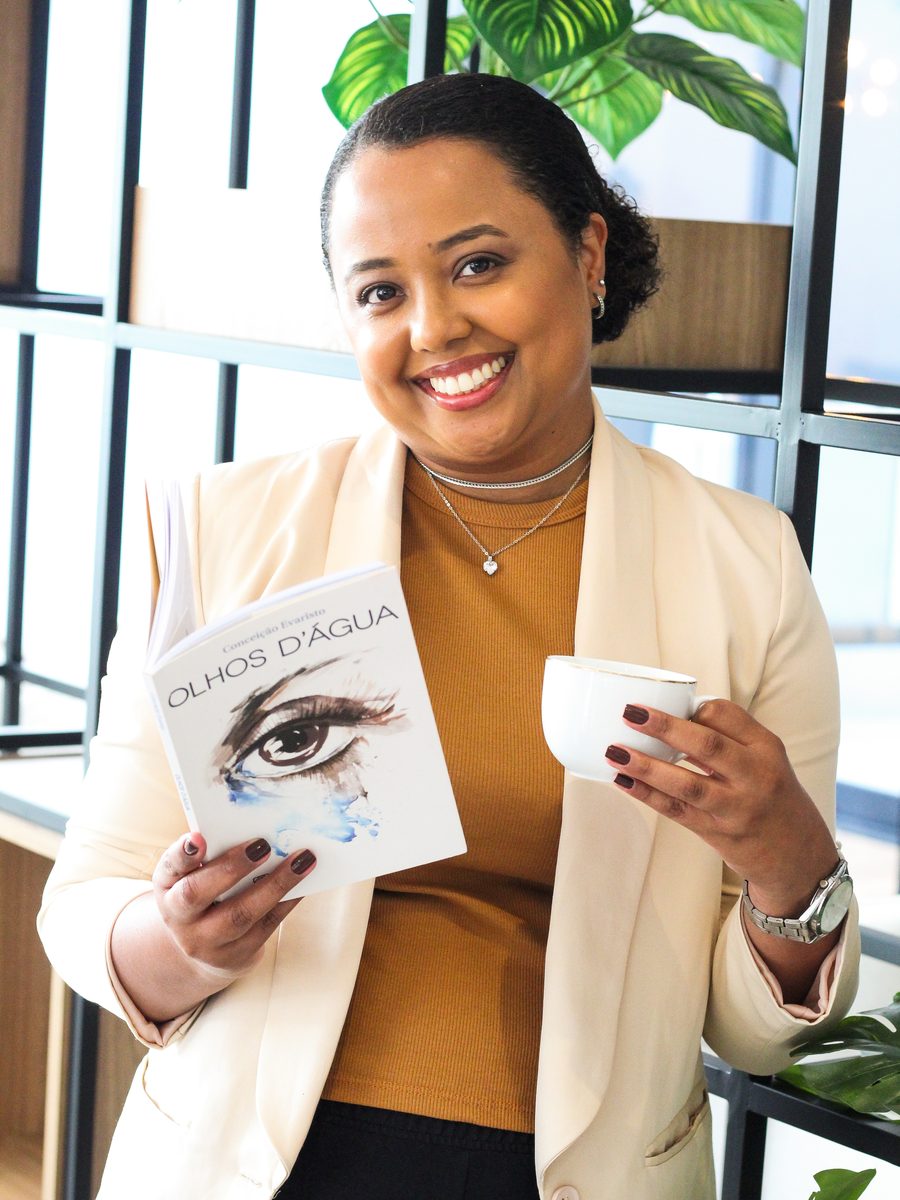 Thaís Marcelino, psicóloga, segurando o livro Olhos d’Água e uma xícara, sorrindo para a câmera.
