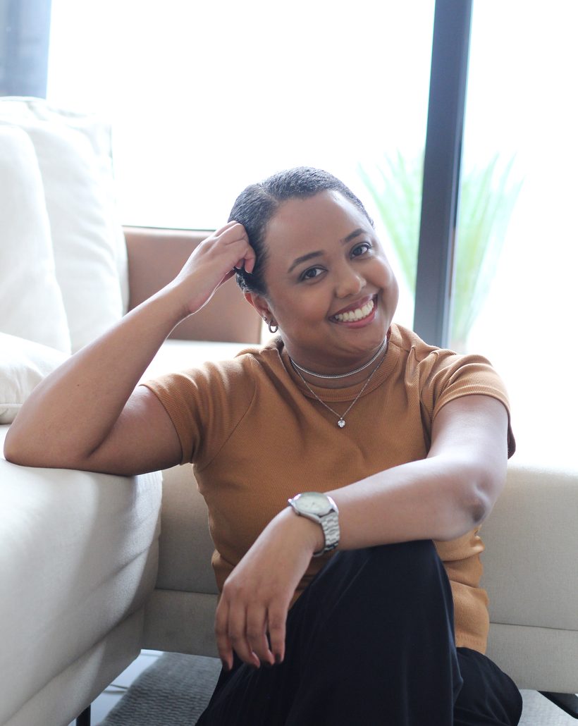 Thaís Marcelino sentada no chão, apoiada em um sofá claro, sorrindo para a câmera.